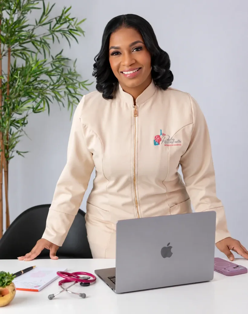 Dra. Rosalba Ruiz Germán con su uniforme médico claro, de pie detrás de su escritorio con una computadora portátil y un estetoscopio