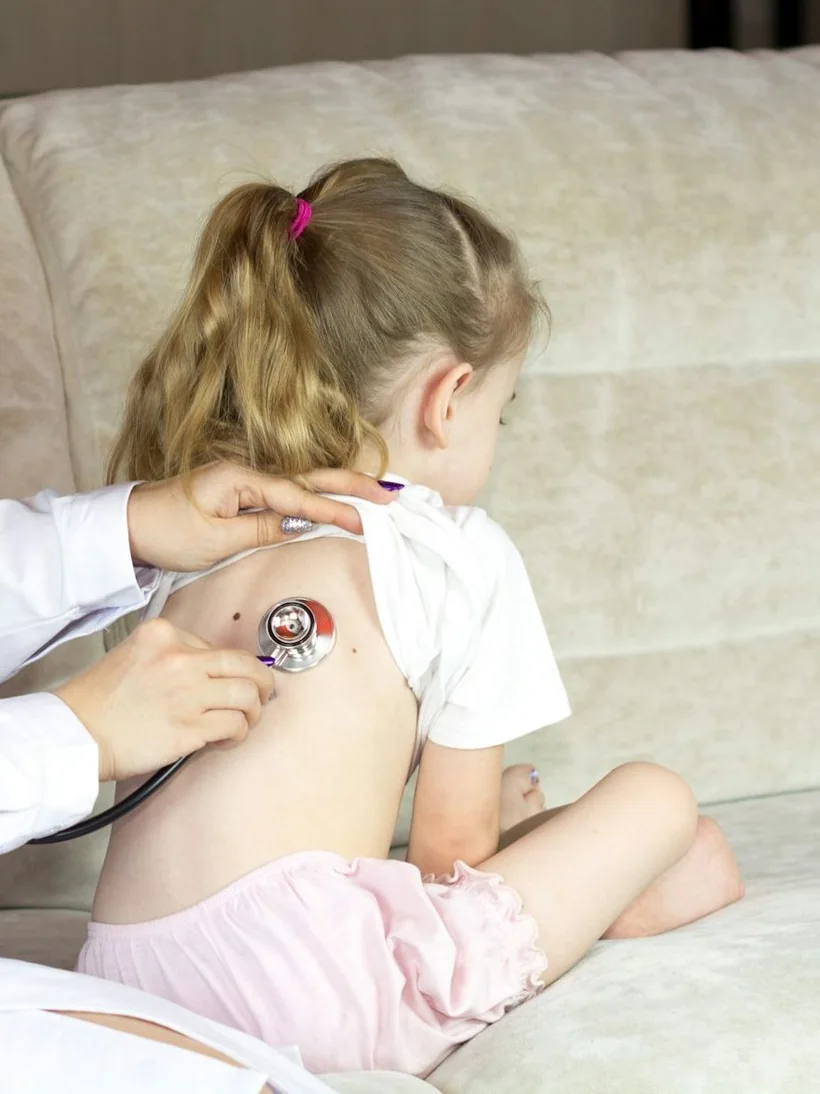 Niña de espaldas sentada en un sofá mientras una profesional escucha su respiración con un estetoscopio